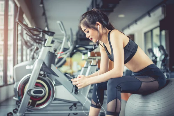 Mulher asiática sentada na bola de fitness e ouvir música no ginásio de treinamento de fitness com telefone inteligente. Relaxe e treino desportivo conceito de treinamento. Tecnologia e entretenimento tema — Fotografia de Stock