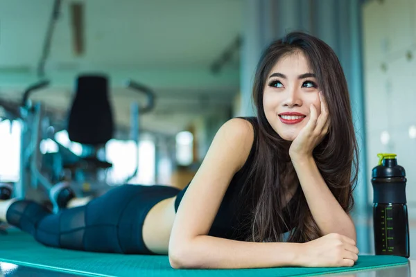 Mujer deportiva tumbada y relajada en la esterilla de yoga en el gimnasio de fitness con equipo deportivo de fondo. Concepto de ejercicio de entrenamiento de belleza y entrenamiento. Cuerpo construir y tema de fuerza . — Foto de Stock