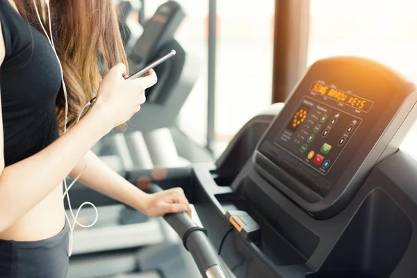 Femeie asiatică care utilizează telefonul inteligent atunci când antrenament sau antrenament de forță la sala de fitness pe banda de alergare. Conceptul de relaxare și tehnologie. Exerciții sportive și teme de îngrijire a sănătății. Stare de spirit fericită și confortabilă . — Fotografie, imagine de stoc