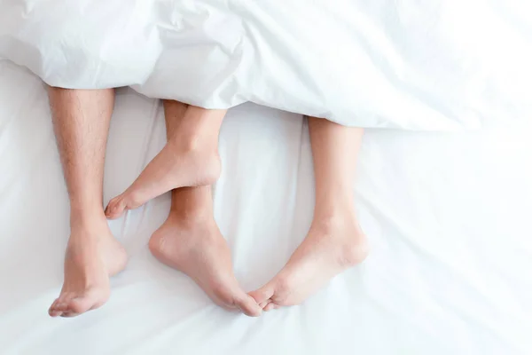 Closeup feet of couple on the bed. Man and woman lovers make love under the blanket or bed sheet. Sex on vacation theme. Valentine and Honeymoon concept. — Stock Photo, Image
