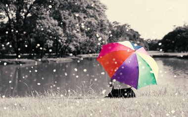 Renkli gökkuşağı şemsiye tutun üzerinde çim sahası, açık Nehri yakınında oturan kadın tarafından doğa ve yağmur, Relax kavramı, güzellik kavramı, yalnız kavramı, seçmeli renk, Sepya dramatik sesi dolu
