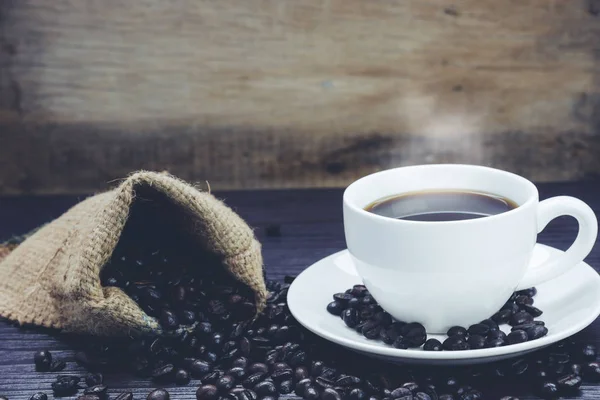 Vista lateral de la taza de café con humo y granos de café en saco sobre mesa de madera, Bebidas y concepto de relax, para publicidad — Foto de Stock