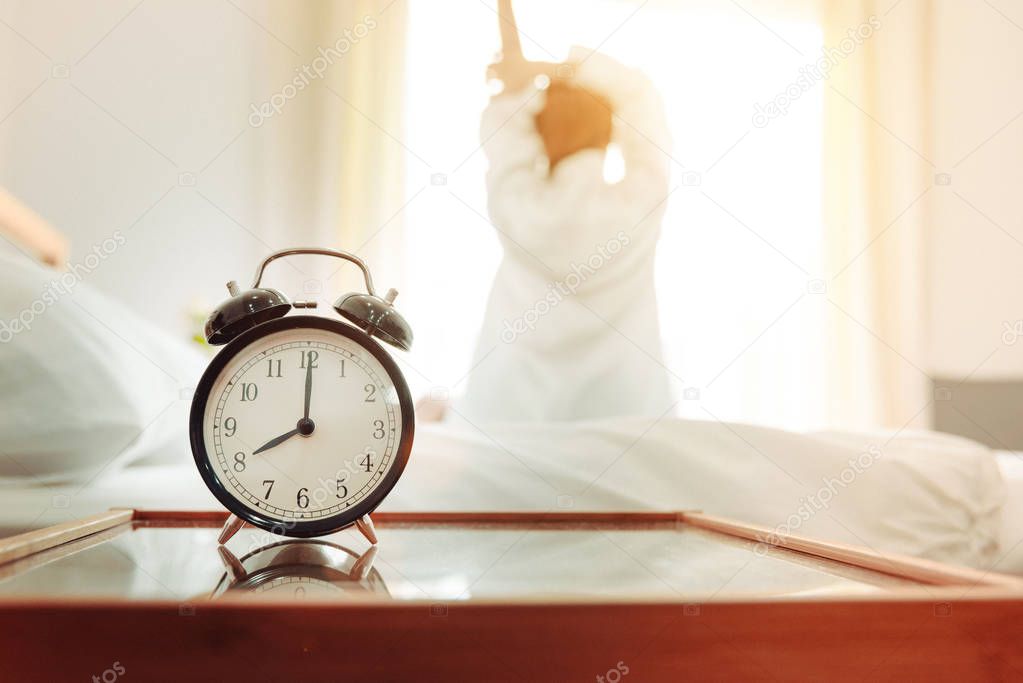 Back view of woman stretching in morning after waking up on bed near window with alarm clock. Holiday and Relax concept. Lazy day and Working day concept. Office woman and worker in daily life theme