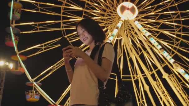 幸せな美しさアジアの女性は観覧車に乗る背景と遊園地の背景で友人とチャットスマートフォンを見ています 人々のライフスタイルとエンターテイメント 旅行と休暇 — ストック動画