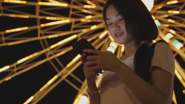快乐美丽的亚洲女人看着智能手机 和朋友在游乐园里聊天 有着摩天轮的背景 人们的生活方式和娱乐 旅行和假日假期 — 图库视频影像