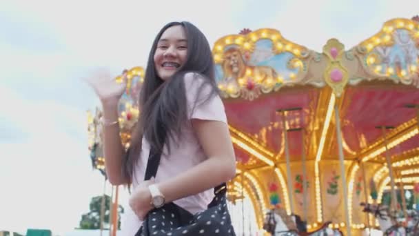 遊園地でカルーセルを楽しむ幸せなアジアの女性 カメラを見て手を振ってください 人々のライフスタイルとエンターテイメント 旅行や休日の概念 楽しい公園カーニバルのテーマの背景 — ストック動画