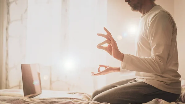 power of meditation, man sitting on the bed in yoga rock position, practicing online at home, details of the hands in surya Mudra e gyan Mudra with filter effect of ray of light