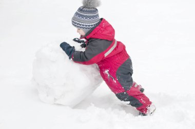 A child sculpts a snowman in the winter on the street, a boy in a red overall clipart