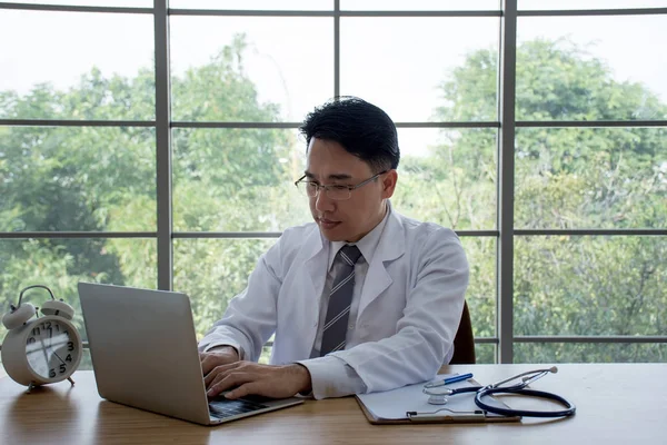 Handsome Asian doctor with laptop and clock on the desk working at a hospital.