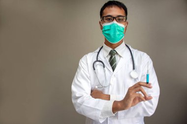 Asyalı doktor kolları tıbbi maskeyle çaprazlanmış gri arka planda bir şırınga tutuyor.