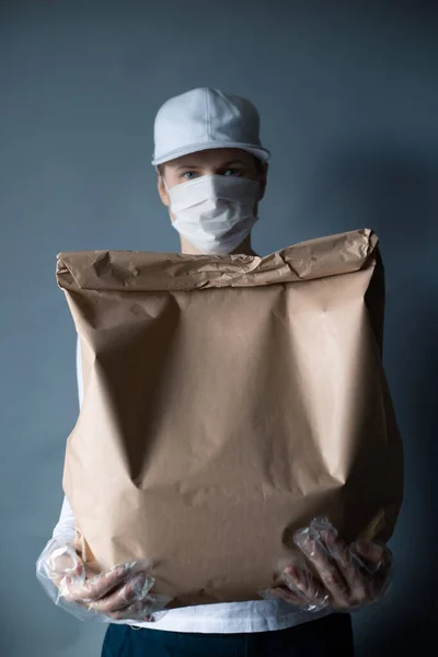 Safe food or goods delivery. Young courier delivering grocery brown eco paper bag order to the home of customer with mask and gloves during the coronavirus pandemic. White clothes gray background copy space for text