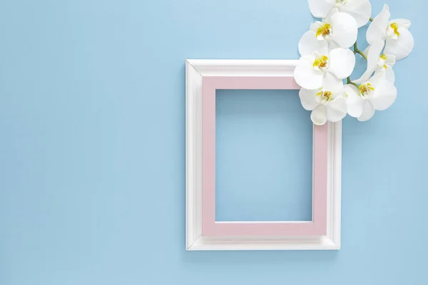 Empty frame and flowers orchid flat lay on blue pastel background with copy space. Minimal concept.