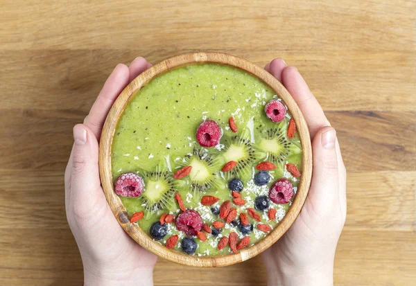 Woman\'s hands holding smoothie bowl. Breakfast kiwi smoothie bowl with with raspberries, blueberries, Goji berries and coconut flakes. Top view.