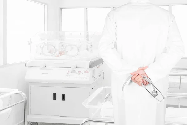 Rear view image of doctors with stethoscope looking at newborn baby covered inside incubator in hospital post-delivery room — Stock Photo, Image
