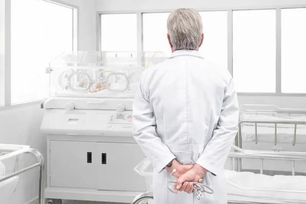 Senior doctor with stethoscope looking at newborn baby covered inside incubator in post delivery room