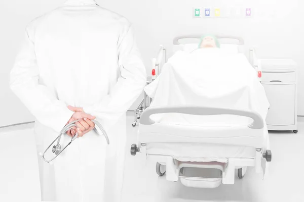 Close-up rear view image of doctors with stethoscope pose arms crossed behind back looking at patient bed in hospital room