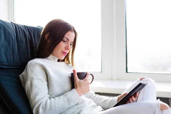 Nainen istuu ikkunan ääressä e-kirjan kanssa. — kuvapankkivalokuva