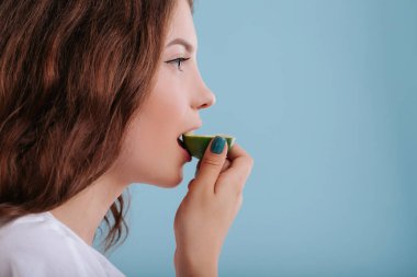Kız limon yiyor, surat asıyor, stüdyoda, mavi arka planda izole edilmiş, kopyalama alanı, kapanış, profil görünümü
