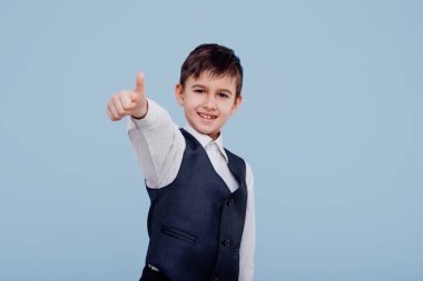 Tişörtlü ve yelekli mutlu küçük çocuk parmağıyla işaret ediyor.