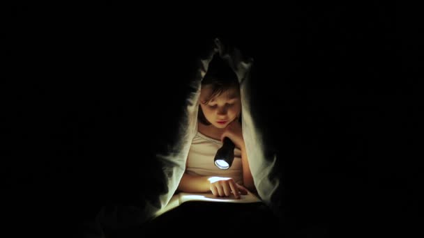A kislány egy könyvet olvas egy takaró alatt egy zseblámpával. — Stock videók
