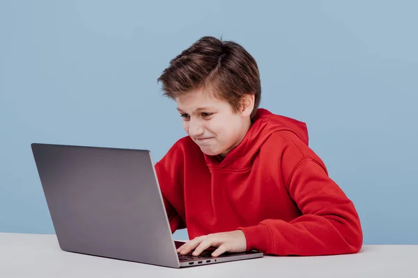 Нервный мальчик в красной толстовке, печатающий на ноутбуке — стоковое фото