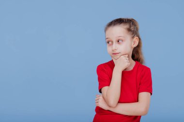 Kırmızı tişörtlü çocuk çeneye dokunuyor ve başka yere bakıyor.