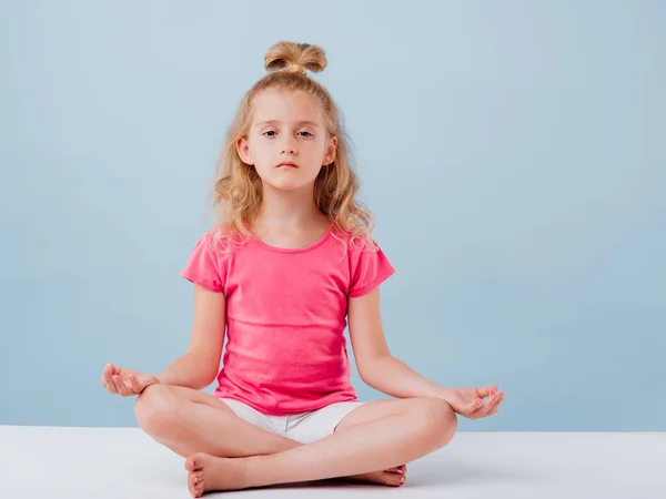 Ein kleines kaukasisches Mädchen sitzt in Yoga-Haltung. isoliert auf blau — Stockfoto