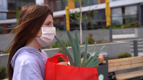 Mujer guapa joven caucásica, máscara médica caminando por la calle y llevando paquete rojo — Vídeos de Stock