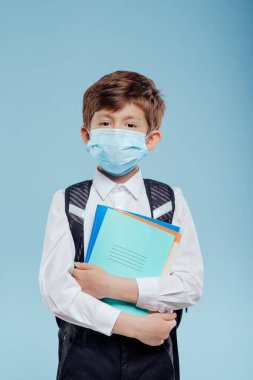 Tıbbi maskeli ve sırt çantalı çocuğun elinde defterler ve kitaplar var.