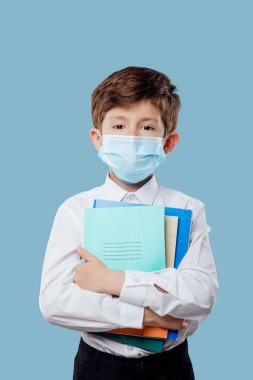 Tıbbi maskeli küçük çocuğun elinde defterler ve kitaplar var.