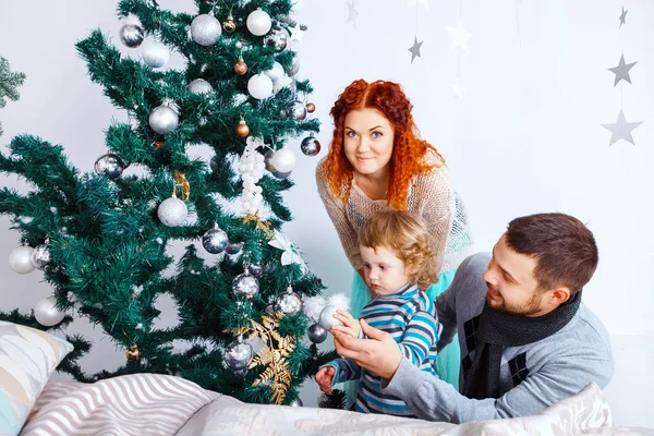 Navidad feliz familia de tres personas decorando abeto con cajas de regalo sobre fondo de dormitorio blanco — Foto de Stock