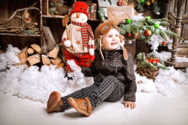 Braun deri ceket, pantolon ve bot üzerinde ahşap dekorasyon kar ile oyunlarında Noel poz pilot şapkası ile küçük iki yaşında bir çocuk giymiş