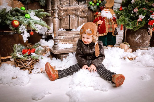 Braun deri ceket, pantolon ve bot üzerinde ahşap dekorasyon kar ile oyunlarında Noel poz pilot şapkası ile küçük iki yaşında bir çocuk giymiş