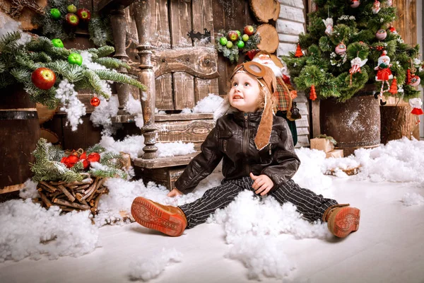 Braun deri ceket, pantolon ve bot üzerinde ahşap dekorasyon kar ile oyunlarında Noel poz pilot şapkası ile küçük iki yaşında bir çocuk giymiş