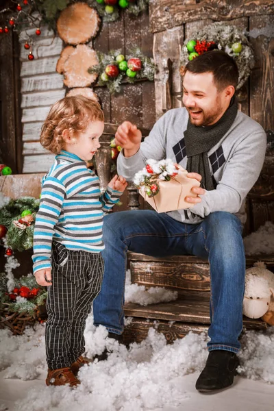 Feliz padre le regala un regalo de Navidad a su hijo en decoraciones con abeto con cajas de regalo y fondo de madera — Foto de Stock