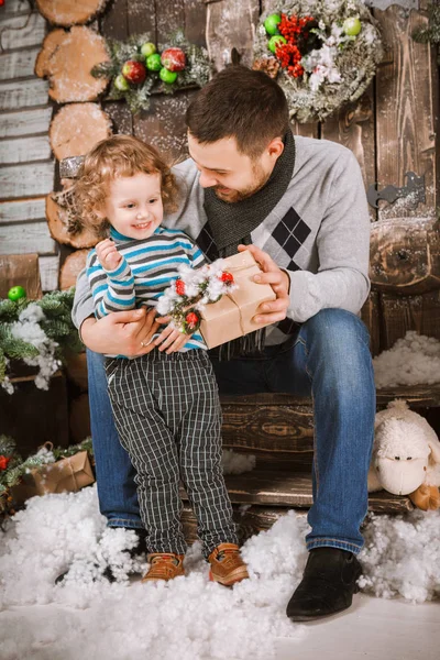 Feliz padre le regala un regalo de Navidad a su hijo en decoraciones con abeto con cajas de regalo y fondo de madera — Foto de Stock