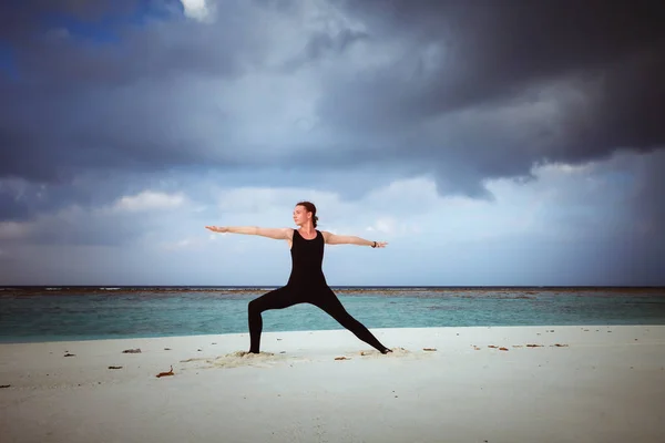 young girl standing in yoga warrior asana on dreamlike beach on summer vacation waering black jumsuit in Maldives