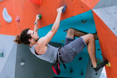 Kıvırcık saçlı, at kuyruklu bir adamın şehir konsepti şehirde talk pudrası kullanan, sigorta ekipmanları olmadan tırmanmak için çanta takan yapay kırmızı ve mavi tırmanma duvarında antrenman yapmaktır.