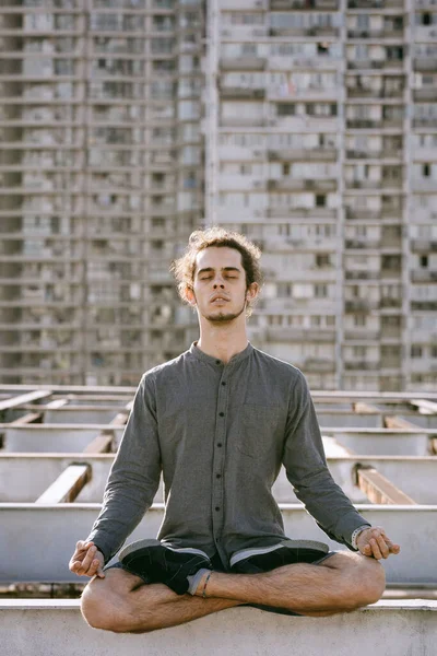 Meditating young european man sitting with eyes closed in lotus pose on blurry urban residential building. Yoga concept — Stock Photo, Image