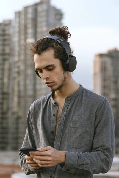 Man sitting on the rooftop and listening music application in wireless black headphones and checking his smartphone