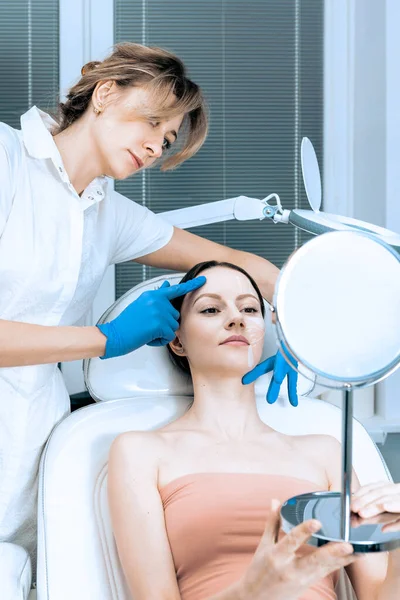 Mujer joven en el servicio de cosmetología de la clínica de belleza sentada en silla médica mientras que la doctora examina la piel con espejo. — Foto de Stock