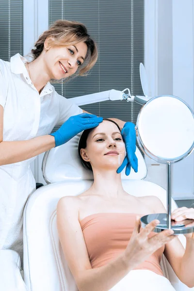 Mujer joven en el servicio de cosmetología de la clínica de belleza sentada en silla médica mientras que la doctora examina la piel con espejo. — Foto de Stock
