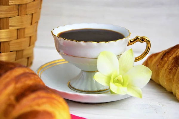 Una taza de café con café expreso caliente en un platillo, croissant, cesta del artículo, flor sobre un fondo pintado de madera — Foto de Stock