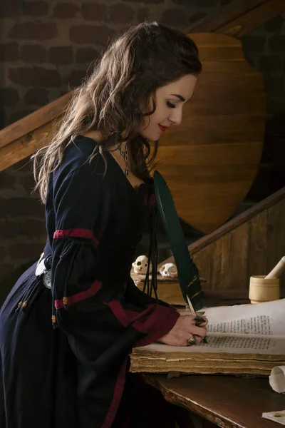 Mujer medieval en un vestido cerca del escritorio con una pluma en la mano. Sobre una mesa yacía el viejo libro y otros elementos mágicos. Brujas del gabinete. Foto de fantasía —  Fotos de Stock