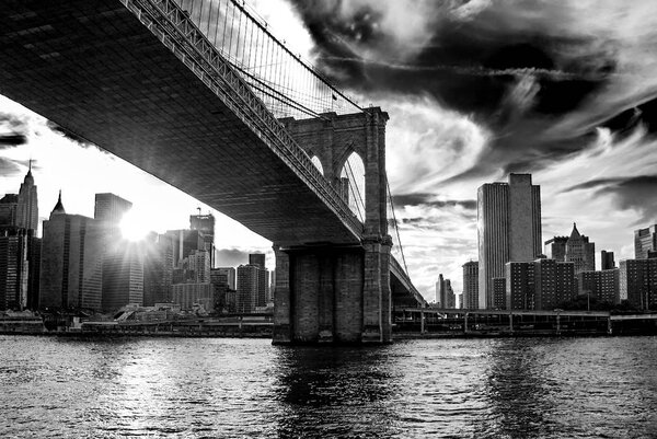 Famous Brooklyn Bridge in New York City, USA with financial district, downtown Manhattan in background. East River and beautiful sunset reflection. Black and white contrast photography, low angle view