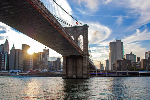 Famous Brooklyn Bridge in New York City, USA with financial district, downtown Manhattan in background. East River and beautiful sunset reflection. Low angle view