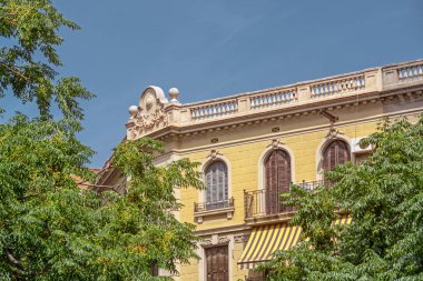 Beautiful building facade through the green trees lit by sunlight, Barcelona, Spain