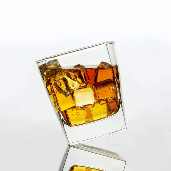 Slanted transparent glass of gold whiskey with pieces of ice against the white background with reflection. Dynamic conceptual scene with alcohol drink