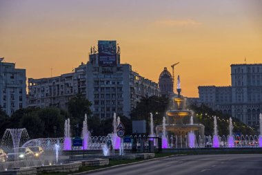 Bucharest, Romania - August 06, 2019: Illuminated colorful fountains in Unirii Square with buildings silhouettes on beautiful sunset sky background