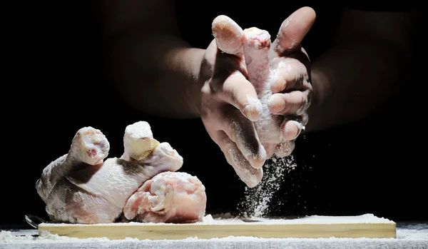 O chef na cozinha prepara frango, embrulhando a perna de frango em farinha. poeira, ação e movimentos na farinha. no fundo preto — Fotografia de Stock